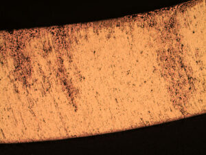 A low magnification photomicrograph of a cross-section through the fitting nipple tube. The un-attacked brass is observed as a bright yellow color while the copper-colored areas are indicative of dezincification. The dezincification attack had penetrated 100% through the tube wall.
